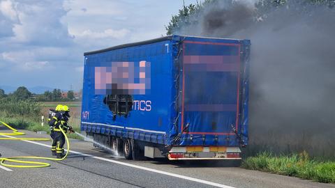 Das Bild zeigt einen blauen Lkw-Anhänger mit einem Loch auf der linken Seite. Zwei Feuerwehrmänner richten einen Löschschlauch auf die Reifen des Lkw.