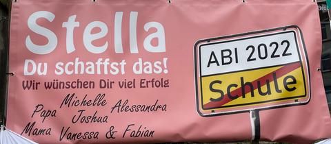 Auf einem rosafarbenen Plakat steht in weißer Schrift "Stella, du schaffst das!" Daneben ein Straßenschild mit der Aufschrift "Abi 2022".