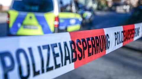 Absperrband mit "Polizeiabsperrung" im Vordergrund des Bildes. Im Hintergund, unscharf, ein Polizeiauto.