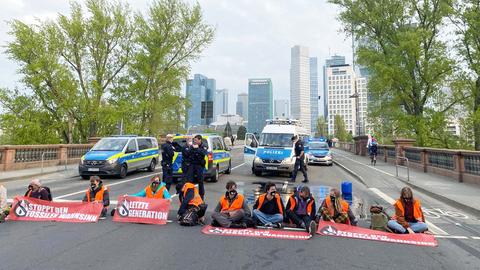 Aktivisten von "Letzte Generation" sitzen auf der Fahrbahn, die zur Untermainbrücke führt. Hinter ihnen Polizisten und Polizeiautos.