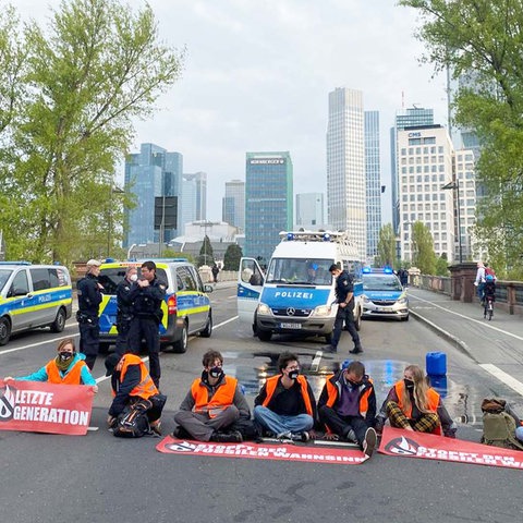 Aktivisten von "Letzte Generation" sitzen auf der Fahrbahn, die zur Untermainbrücke führt. Hinter ihnen Polizisten und Polizeiautos.