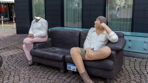 Das Bild zeigt eine Skulptur aus Beton, die auf einer Bank sitzt und der der Kopf abgeschlagen wurde. 
