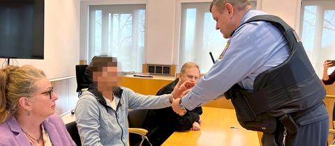 Das Foto zeigt den Angeklagten, sein Gesicht ist verpixelt, wie er am Tisch sitzt und die Handschellen von einem Justizbeamten abgenommen bekomt´mt.