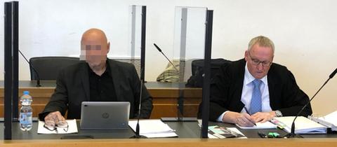 Links sitzt hinter einem Laptop der Angeklagte an einem Tisch im Gerichtssaal, rechts neben ihm mit blauer Krawatte sein Anwalt.