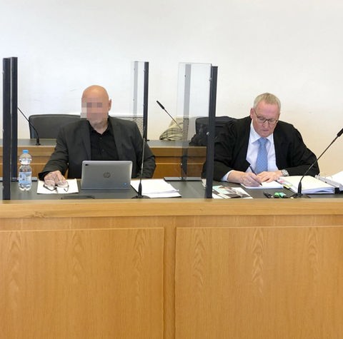 Links sitzt hinter einem Laptop der Angeklagte an einem Tisch im Gerichtssaal, rechts neben ihm mit blauer Krawatte sein Anwalt.