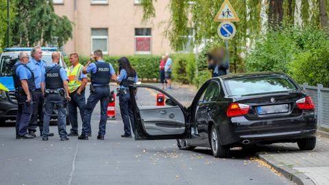 Blick in eine Straße. Rechts, halb auf dem Bürgersteig, steht ein beschädigtes Auto mit geöffneter Tür. Links im Bild, auf der Straße, stehen mehrere Einsatzkräfte der Polizei.