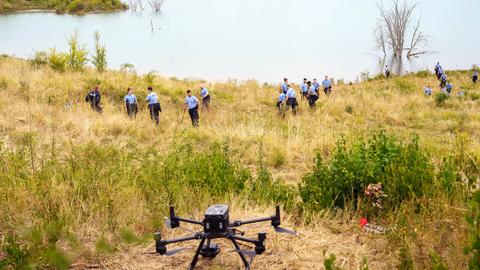 Im Bildvordergrund eine Drohne auf dem Boden einer Wiese. Im Bildhintergund streifen viele Polizisten durch die Wiese am Rande eines Sees.