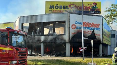 Ausgebranntes Fabrikgebäude, das ein Möbelhaus ist, davor Feuerwehrwagen, darüber Rauch