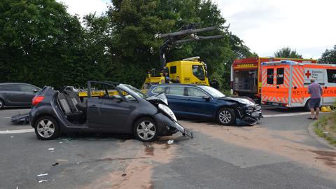 Zwei beschädigte Autos stehen auf einer Landstraße, daneben ein Abschleppwagen, ein Feuerwehrauto und ein Krankenwagen.