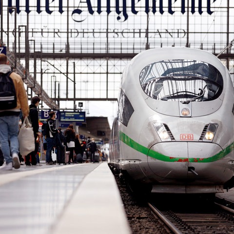 Passagiere mit Gepäck auf einem Bahnsteig an einem Gleis, auf dem ein ICE steht.