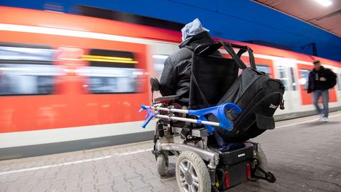Ein Mann in einem Rollstuhl am Bahnsteig. Ein Zug fährt ein, weshalb er unscharf im Hintergrund ist.