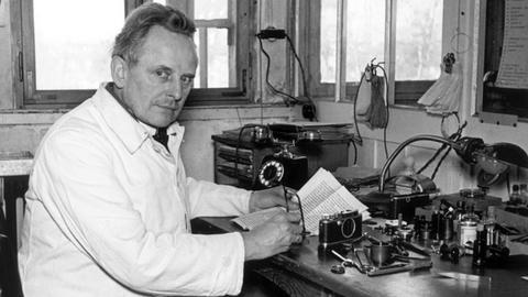 Schwarz-Weiß-Foto. Ein Mann mittleren Alters sitzt im Kittel an einem Schreibtisch mit Fotoapparaten.