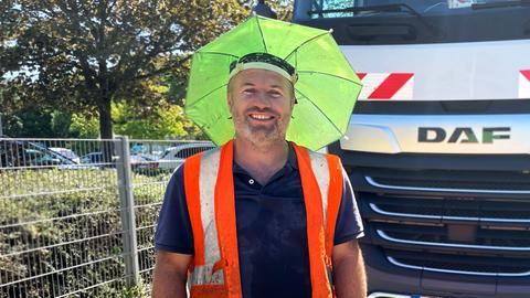 Ein Mann mit einem gelben Kopfsonnenschutz, der aussieht wie ein Regenschirm, der direkt am Kopf befestigt wird, steht vor einem LKW und lacht in die Kamera.