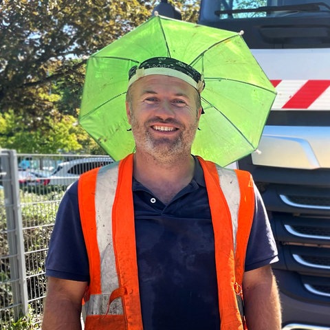 Ein Mann mit einem gelben Kopfsonnenschutz, der aussieht wie ein Regenschirm, der direkt am Kopf befestigt wird, steht vor einem LKW und lacht in die Kamera.