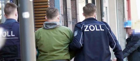 Ein Mann in grüner Jacke wird von zwei Beamten in Jacken mit der Aufschrift "Zoll" untergehakt und abgeführt. Sie laufen auf einem Bürgersteig und sind von hinten zu sehen.
