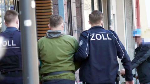 Ein Mann in grüner Jacke wird von zwei Beamten in Jacken mit der Aufschrift "Zoll" untergehakt und abgeführt. Sie laufen auf einem Bürgersteig und sind von hinten zu sehen.