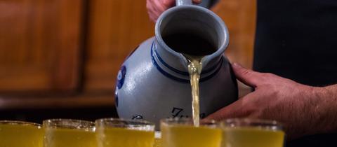 Hände halten einen "Apfelwein-Bembel" und gießen eine hellgelbe Flüssigkeit in viele Gläser.