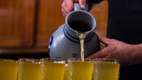 Hände halten einen "Apfelwein-Bembel" und gießen eine hellgelbe Flüssigkeit in viele Gläser.