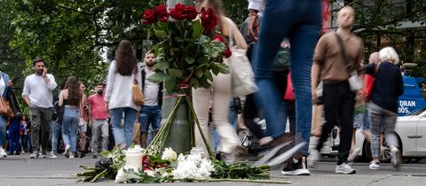 Passanten gehen auf einer belebten Straße an Blumen vorbei, die an einem Ampelmast niedergelegt wurden.