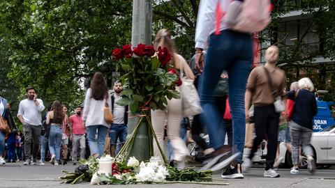 Passanten gehen auf einer belebten Straße an Blumen vorbei, die an einem Ampelmast niedergelegt wurden.