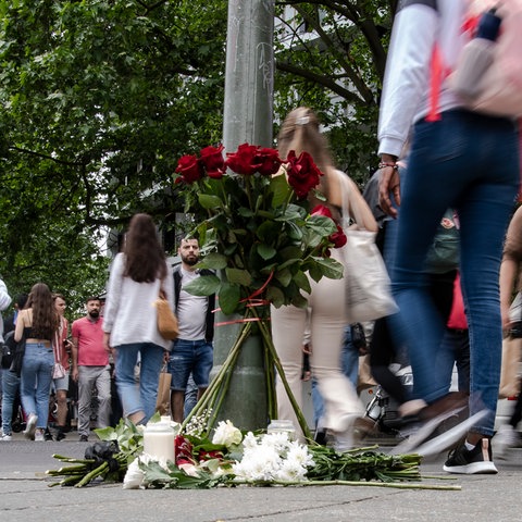 Passanten gehen auf einer belebten Straße an Blumen vorbei, die an einem Ampelmast niedergelegt wurden.