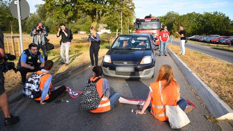 Drei Menschen in Warnwesten sitzen auf einer Straße, dahinter stauen sich Autos.