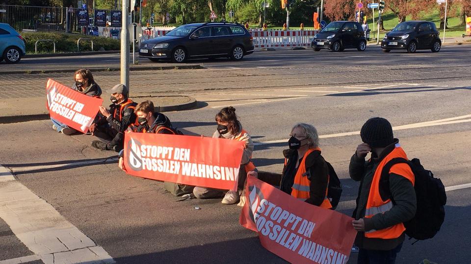 Sechs Menschen mit Warnwesten sitzen auf einer Straße. Sie halten Transparente mit der Aufschrift "Stoppt den fossilen Wahnsinn".