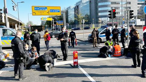 Auf einer mehrspurigen Straße stehen mehrere Polizistinnen und Polizisten. Auf der Straße sitzen Menschen in Warnwesten.