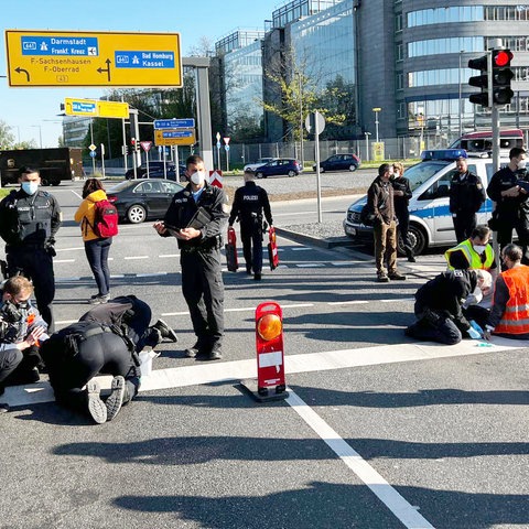 Auf einer mehrspurigen Straße stehen mehrere Polizistinnen und Polizisten. Auf der Straße sitzen Menschen in Warnwesten.