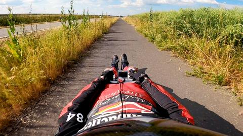 Foto eines Mannes in Rennsportkleidung, wie er auf einem kleinen Bobbycar liegt und in Richtung eines Radweges schaut. Die Kamera ist auf dem Helm des Fahrers angebracht, so dass das Bild den Körper abwärts zeigt vor dem Hintergund eines Radweges der auf den Horizont zuläuft.