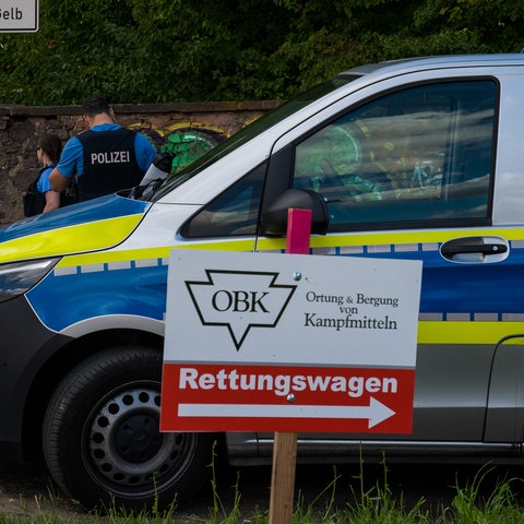 Auf einem Schild steht "Ortung und Bergung von Kampfmitteln", daneben ein Polizeiauto.