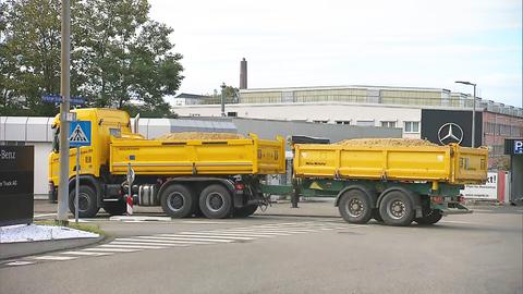 Ein mit Sand beladener Lkw steht vor dem Tor eines Werksgeländes. Im Hintergrund ist ein Schild mit einem Stern, dem Logo von Mercedes-Benz, zu sehen.
