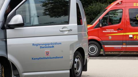 Das Bild zeigt eine silberne Autotür mit der Aufschrift "Regierungspräsidium Darmstadt - Kampfmittelräumdienst" und dahinter ein rotes Auto der Feuerwehr Rüdesheim.