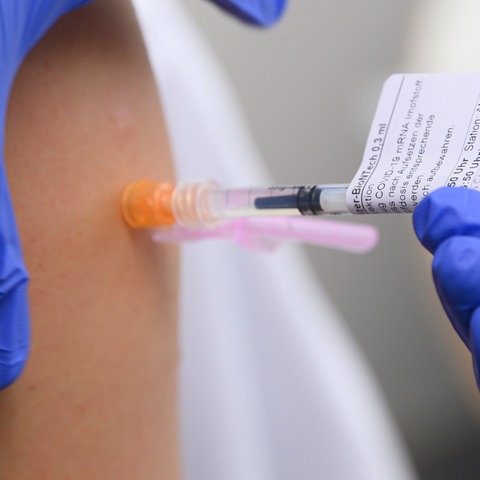 Zwei Hände mit blauen Schutzhandschuhen führen eine Impfung an einem Oberarm aus.