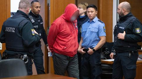 Mann mit roter Kapuzenjacke wird von Justizbeamten in Raum geführt