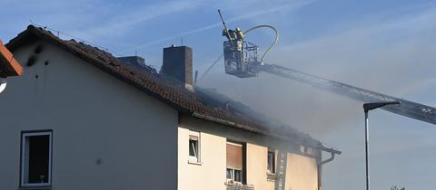 Feuerwehrleute stehen um ein Wohnhaus mit verkohltem Dachstuhl herum, aus dem Rauch quilllt.
