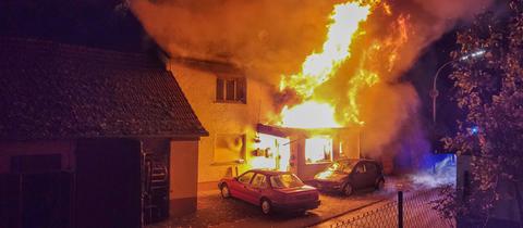 Zwei Autos stehen vor einem Haus, aus dessen Eingang und dessen Fenstern meterhohe Flammen schlagen.