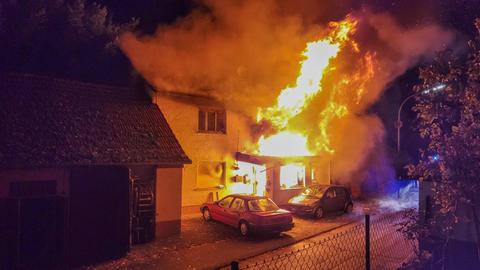 Zwei Autos stehen vor einem Haus, aus dessen Eingang und dessen Fenstern meterhohe Flammen schlagen.