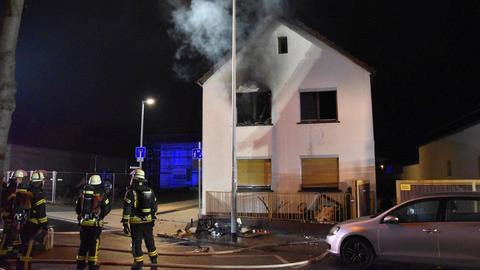 Einsatzkräfte der Feuerwehr bei den Löscharbeiten eines Einfamilienhauses. Aus dem obersten linken Fenster strömt Rauch.