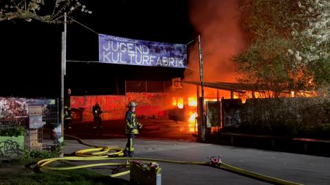 Ein Feuerwehrmann steht vor dem Jugendzentrum, auf dem Autos brennen. Flammen sind im Hintergrund zu sehen. 
