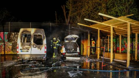 Zwei ausgebrannte Fahrzeuge, ein Kleintransporter und ein SUV, stehen auf einem Parkplatz. Daneben ein Feuerwehrmann mit einem Schlauch.