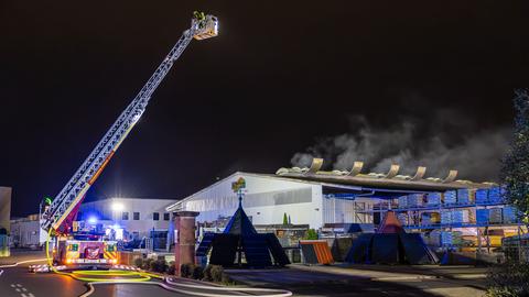 Löscharbeiten an einer Lagerhalle in Hanau - Feuerwehr mit Drehleiter vor Nachthimmel - aus dem Gebäude steigt Rauch auf. 