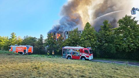 Zwei Feuerwehrautos und -einsatzkräfte stehen vor einem Gebüsch. Dahinter lodern Flammen und steigen Rauchschaden auf.