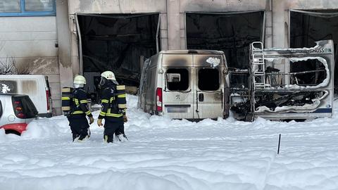 Feuerwehrmänner stehen vor ausgebrannten Autos, der Boden ist mit Löschschaum bedeckt