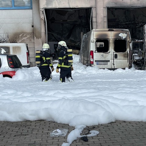 Feuerwehrmänner stehen vor ausgebrannten Autos, der Boden ist mit Löschschaum bedeckt
