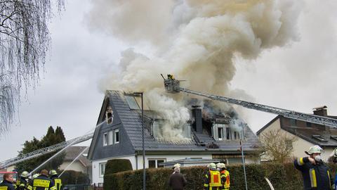 Rauch steigt aus Dach eines Einfamilienhaus hoch, von zwei Seiten sind Feuerwehrleitern und Löschwasser zu erkennen, auf der Straße stehen Feuerwehrkräfte.