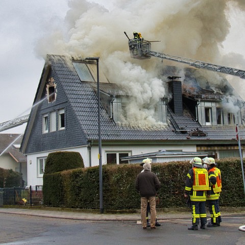 Rauch steigt aus Dach eines Einfamilienhaus hoch, von zwei Seiten sind Feuerwehrleitern und Löschwasser zu erkennen, auf der Straße stehen Feuerwehrkräfte.