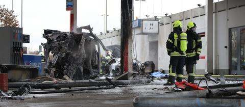 Zwei Feuerwehrleute stehen an der Tankstelle neben dem ausgebrannten Autowrack