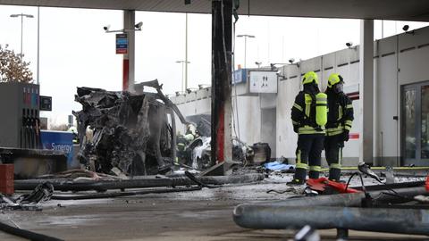 Zwei Feuerwehrleute stehen an der Tankstelle neben dem ausgebrannten Autowrack