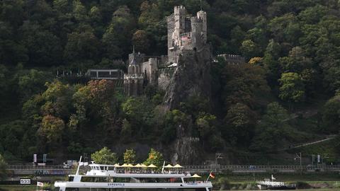 Eine Burg thront über einem Fluss, auf dem ein Ausflugsschiff fährt.